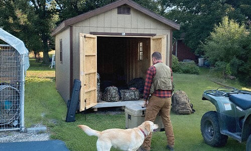 Hunter organizing gear inside a Dakota Storage shed with dog and ATV nearby, showcasing secure outdoor storage solution