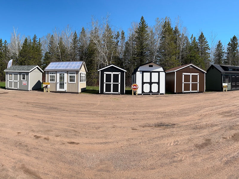Row of display sheds at Dakota Storage Buildings lot in Ashland, Wisconsin, with various shed styles and colors shown on a gravel lot with a tree line in the background