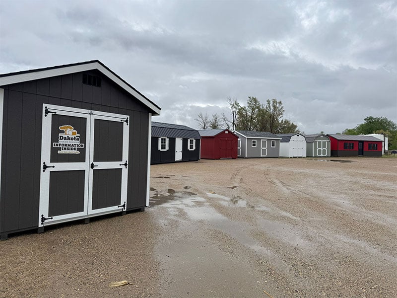 Dakota Storage Buildings display lot featuring a white and black shed with multiple windows and a nearby sign that reads 